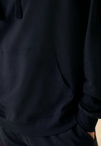 Granatowy bluza z kapturem z przednią kieszenią, teksturowana tkanina i sznurek przy kapturze. Posiada ściągacze przy mankietach i u dołu oraz luźny krój.