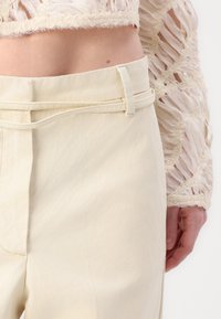 Beige hochgeschnittene Hose mit flacher Vorderseite, seitlichen Taschen und einem Stoffgürtel-Detail. Glatte Textur mit subtilen Stichakzenten.