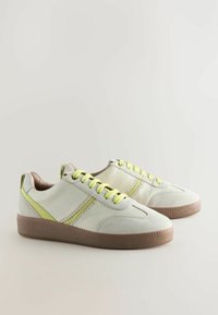 White sneakers with yellow laces and side accents. Features a textured sole, smooth leather, and stitched detailing along the sides.