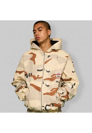 Mężczyzna w beżowo-brązowej, moro, zapinanej na zamek kurtce z kapturem, z rękami w kieszeniach, stojący na tle jednolitego szarego tła.