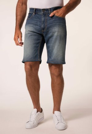 Mand iført falmede blå denimshorts og hvide sneakers, stående med den ene hånd i lommen mod en ensfarvet baggrund.