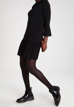 Frau trägt ein schwarzes Kleid mit ausgestellten Ärmeln, schwarze gepunktete Strumpfhosen und glänzende schwarze Stiefeletten und geht vor einem einfarbigen Hintergrund.