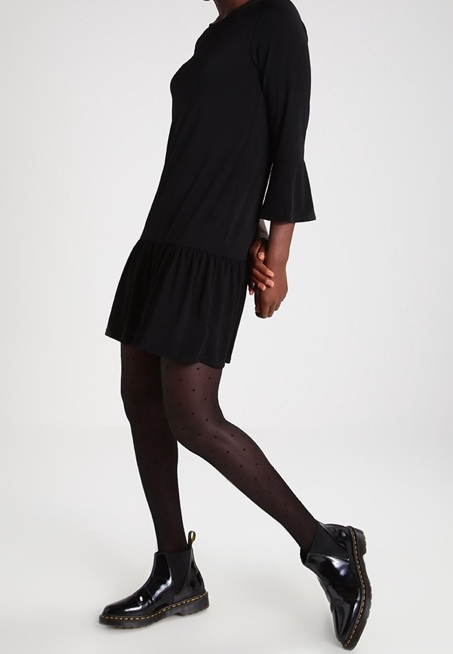 Femme portant une robe noire avec des manches évasées, des collants noirs à pois et des bottines noires brillantes, marchant devant un fond uni.