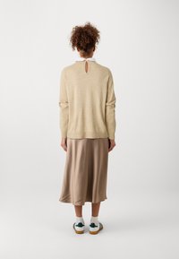 Beżowy sweter wykonany na drutach z detalem w kształcie dziurki na plecach, zestawiony z satynową beżową spódnicą, noszony z białymi trampkami, prezentujący swobodny, warstwowy wygląd.