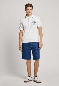Balta polo krekla ar tumši zilu apkakles detaļu un logotipu, apvienots ar zilu džinsu šortiem. Modelis valkā baltas kedas ar melniem akcentiem.