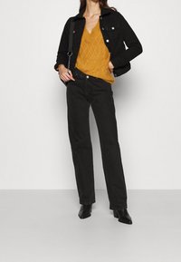 Zwarte denimjack, mosterdgeel gebreid sweater met een ruitpatroon, zwarte hoge spijkerbroek en zwarte hakken.