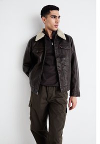 Donkerbruine leren jas met een schapenvachtkraag, twee bor pockets en een getailleerd silhouet, gedragen over een donker shirt en olijfgroene cargo pants.