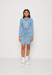 Denimdress i ljusblått med långa ärmar, dragkedja fram och matchande bälte i midjan. Stylad med vita sneakers och ankelsockar.