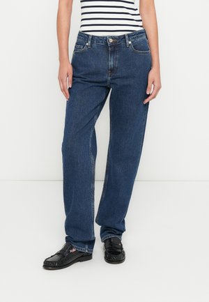 Personne portant un jean bleu taille haute à jambes droites, des mocassins noirs et un haut sans manches rayé blanc et noir.