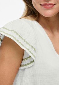 Blusa blanca con tela texturizada, presenta un borde de ondas en verde oliva a lo largo de las mangas y un sutil diseño de escote en V.