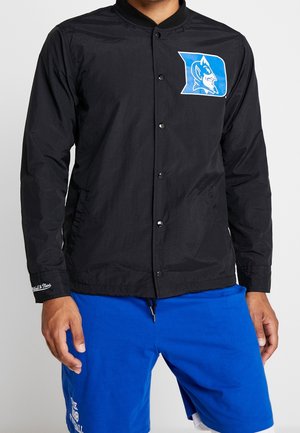 Mand iført en sort jakke med trykknapper med et blåt Duke Blue Devils-logo på brystet og blå atletikshorts.
