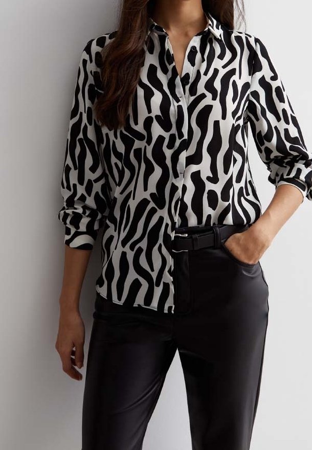 Femme portant un chemisier à manches longues avec un motif abstrait noir et blanc, un pantalon noir taille haute et une ceinture noire, une main dans la poche.