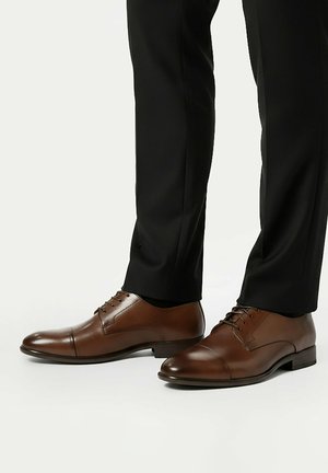 Braune Oxford-Lederschnürschuhe, getragen mit schwarzen Hosen, von der Seite vor weißem Hintergrund gezeigt.