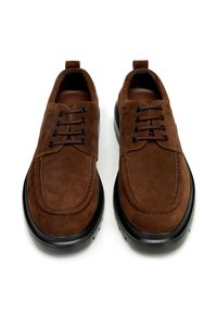 Par de zapatos de ante marrón con cordones, suelas gruesas negras, punteras redondeadas y detalles de costura, vistos desde arriba sobre un fondo blanco.