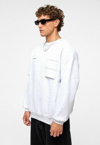 Sweat-shirt oversize gris clair à manches longues, doté d'une poche avant et d'un ourlet côtelé. Texture lisse, porté avec un pantalon noir et des lunettes de soleil.