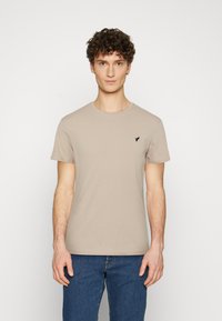 Beige cotton t-shirt with a round neck and short sleeves, featuring a small black bird logo on the left chest. Worn with blue jeans.
