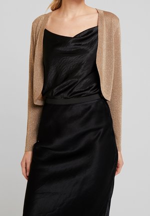 Femme portant un cardigan court doré scintillant sur une robe noire en satin sans manches avec un décolleté asymétrique.