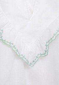 White fabric with delicate green floral embroidery along a ruffled neckline. The texture is soft, and the design features a V-shaped cut.