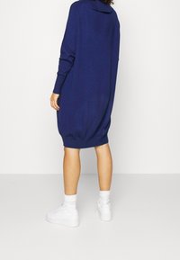 Abito a maglia blu navy, oversize, con maniche lunghe e orlo curvo, realizzato in materiale morbido. Abbinato a sneaker bianche.
