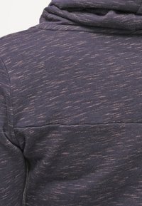 Detailní záběr na fialovou heatherovou látku s horizontálním švem a částí kapuce viditelnou v horní části.