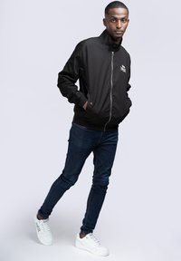 Chaqueta bomber negra con cremallera y cuello alto, combinada con jeans skinny azul oscuro y zapatillas blancas. Diseño simple sin logotipos visibles.
