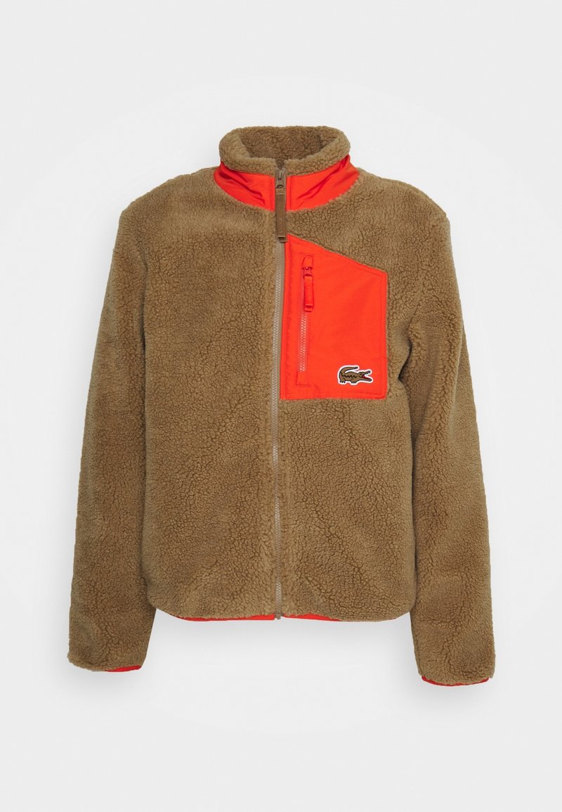 Veste en polaire marron avec un col montant, présentant une poche zippée orange vif et un petit logo sur le côté. Tissu texturé sur l'ensemble.