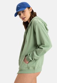 Grön zip-up hoodie med dragsko, framfickor och ribbade ärmslut. I kombination med en blå basebollkeps med vit brodyr.