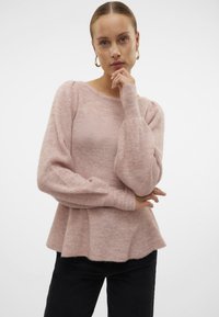 Svetlo roza pleteni pulover z okroglo ovratnikom, napihnjenimi rokavi in razširjenim robom. Tekstura izgleda mehka in udobna.