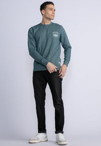 Türkiser Crewneck-Pullover mit Logo auf der Brust, kombiniert mit dunklen Jeans und weißen Sneakern. Weicher Stoff mit gerippten Bündchen und Saum.
