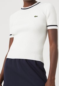 Bílý krátký rukáv s žebrovaným úpletem s námořnicky modrým lemováním u výstřihu a rukávů, s malým zeleným vyšívaným logem na hrudi.