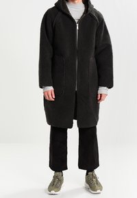 Musta fleece-takki, jossa vetoketjullinen edessä, huppu ja kaksi suurta taskua; päällä harmaa paita ja yhdistetty mustiin puuvillahousuihin ja lenkkareihin.