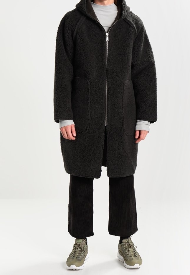 Musta fleece-takki, jossa vetoketjullinen edessä, huppu ja kaksi suurta taskua; päällä harmaa paita ja yhdistetty mustiin puuvillahousuihin ja lenkkareihin.