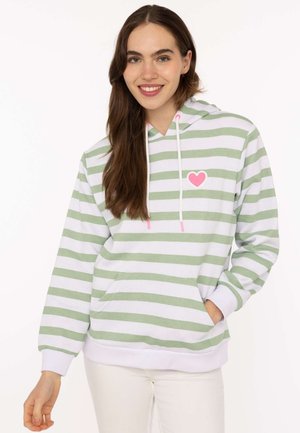 Junge Frau lächelt, trägt einen grün-weiß gestreiften Hoodie mit einem rosa Herz auf der Brust und weiße Hose, steht mit einer Hand in der Tasche.