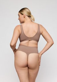 Mujer con cabello rubio recogido en una cola de caballo, vestida con un sujetador beige y una tanga de talle alto, de espaldas a la cámara contra un fondo simple.