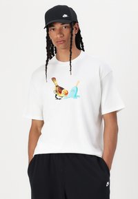 Hvid T-shirt med et farverigt fugl- og ispop-grafik. Modellen bærer en sort kasket. Mørke bukser fuldender outfittet.