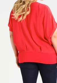 Top rosso a maniche corte con vestibilità morbida e orlo arricciato, abbinato a jeans scuri. Il tessuto appare liscio e leggero con una caduta morbida.