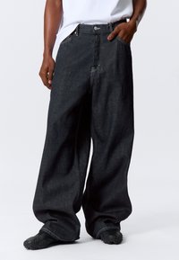 Široke tamne traperice s visokim strukom, s jednim prednjim džepom i kontrastnim šavovima. Nosene s crnim teksturiranim cipelama.