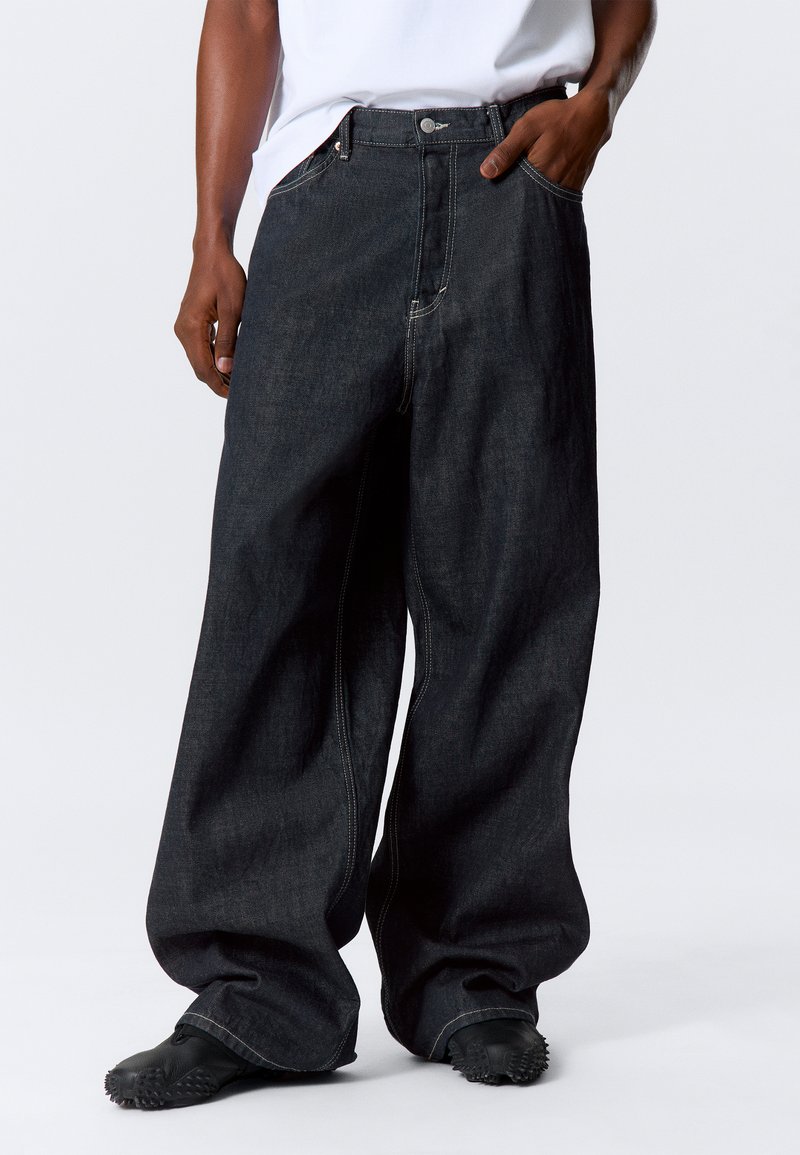 Široke tamne traperice s visokim strukom, s jednim prednjim džepom i kontrastnim šavovima. Nosene s crnim teksturiranim cipelama.