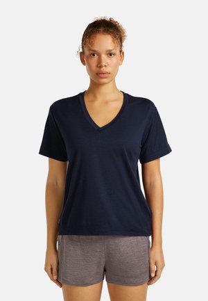 Junge Frau mit lockigem Haar zu einem Dutt gebunden, trägt ein marineblaues V-Ausschnitt T-Shirt mit kurzen Ärmeln und graue Shorts, steht vor einem weißen Hintergrund.