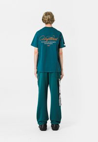 Camiseta de algodón en color verde azulado con mangas cortas y texto en la parte posterior. Combinada con pantalones de corte relajado a juego. Presenta calzado negro texturizado único.