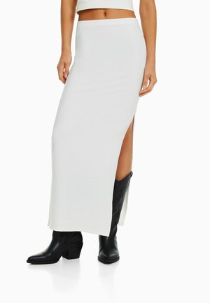 Kvinde iført en hvid maxi-nederdel med høj talje og et lårslids, kombineret med sorte cowboystøvler med hæl, der går til midt på læggen.