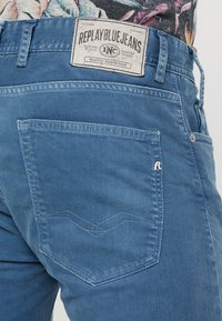 Siniset farkut matalalla vyötärölinjalla, jossa on suorakulmainen takatasku ompeluyksityiskohdalla ja etiketti, jossa lukee "REPLAY BLUE JEANS."