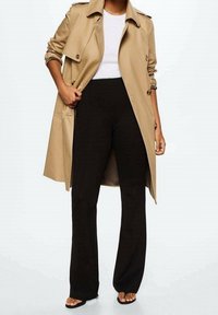 Beżowy płaszcz trench na białej bluzce, połączony z czarnymi spodniami w kształcie dzwonów i czarnymi płaskimi sandałami. Posiada guziki i luźny krój.