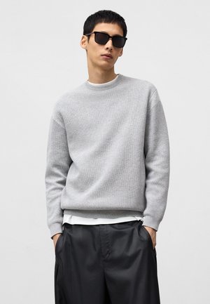 Light gray knitted sweater with a round neckline, ribbed cuffs, layered over a white shirt. Model wears black square sunglasses and loose dark pants.