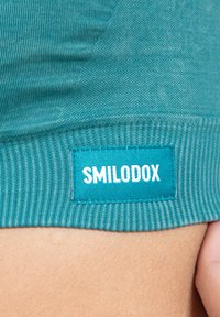 Vihreä neulekangas, jossa on ribbitekstuuri. Ominaisuuksina suorakulmainen etiketti, jossa lukee "SMILODOX" valkoisella sinisellä taustalla. Lähikuvassa.