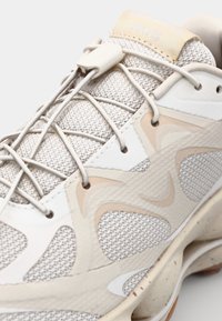 Light beige athletic shoe featuring a mesh upper, tonal accents, and a textured sole. Laces are integrated with a plastic toggle.