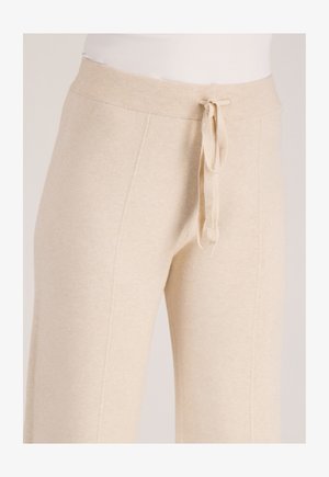 Beige gebreide broek met een geribbelde elastische tailleband en voorstrik, gedragen door een persoon met een wit topje.