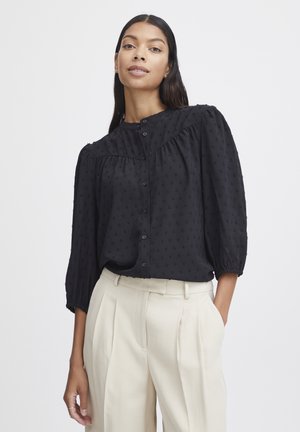 Zwarte blouse van gestructureerde stof, met knoopsluiting en geplooide schouders. Gecombineerd met beige geplooide broeken.