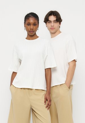 Δύο μοντέλα στέκονται δίπλα δίπλα φορώντας λευκά ριμπ κοντομάνικα τοπ και μπεζ παντελόνια με φαρδιά πόδια σε απλό φόντο.