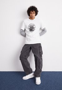 Valkoinen graafinen t-paita, jossa pyöreä musta kuvio, käytetty harmaan pitkähihaisen paidan päällä. Tumma cargo-housut ja valkoiset lenkkitossut täydentävät asun.